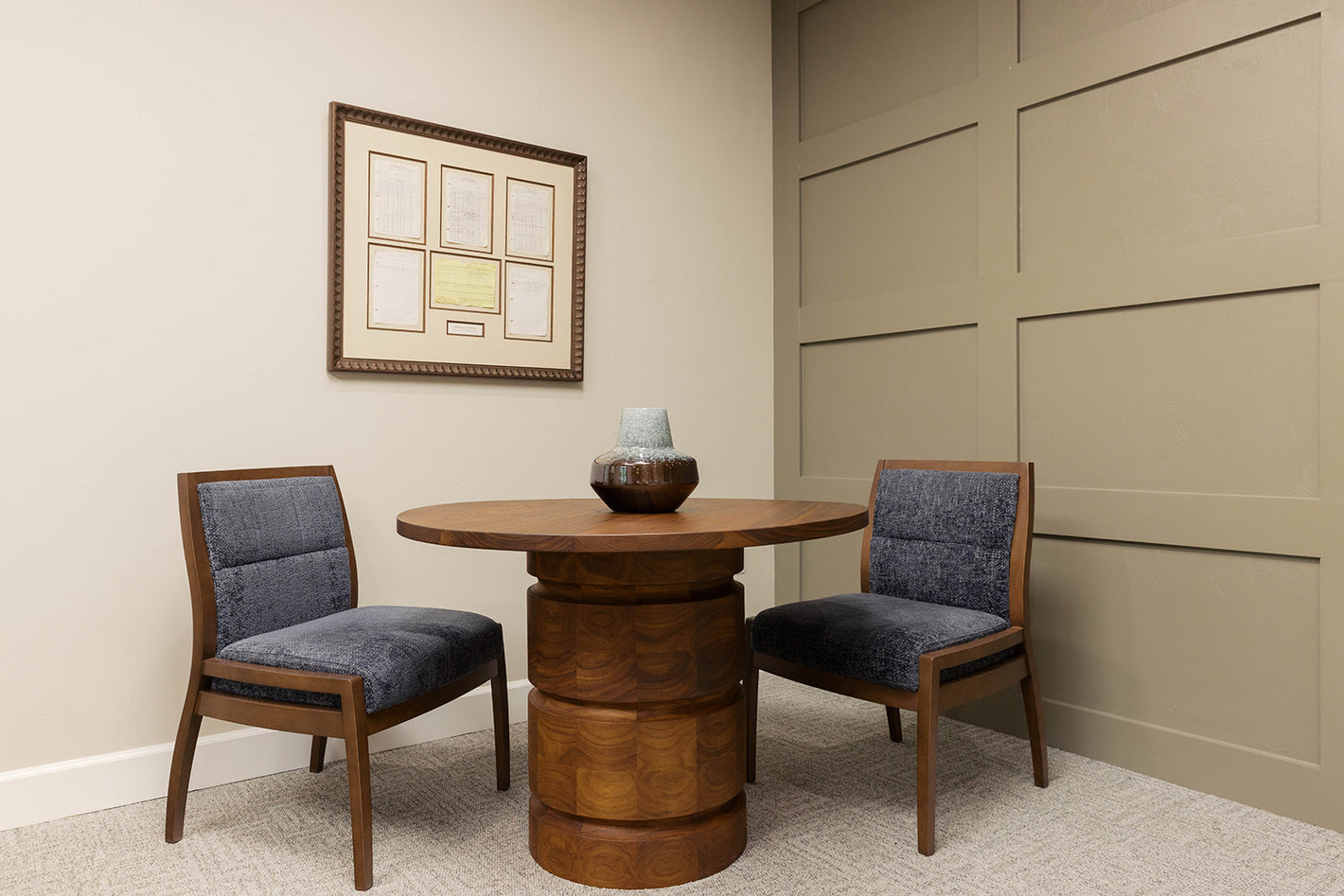A round wooden pedestal table with two matching wooden chairs featuring blue upholstered seats, set against a beige paneled wall and cream wall with a framed certificate on display.