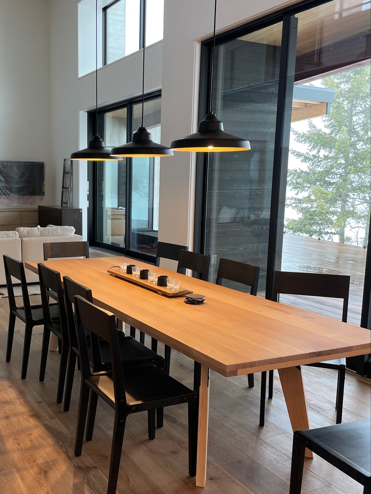 A modern dining room scene with a long light wood dining table surrounded by black chairs, under three black pendant lights. A tan dog is lying on a light rug in the adjacent living area.