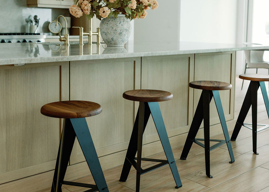 A modern kitchen scene with four bar stools in front of a light wood island, two dark dome pendant lights overhead, and a vase of flowers on the counter.