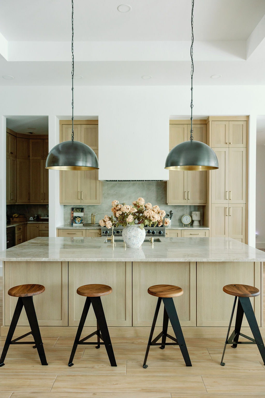 A modern kitchen island with a light wood base and marble countertop, four wooden bar stools with black metal legs, and two dark dome pendant lights hanging above. The background features light wood cabinetry, a stone backsplash, and a vase of pink flowers on the island.